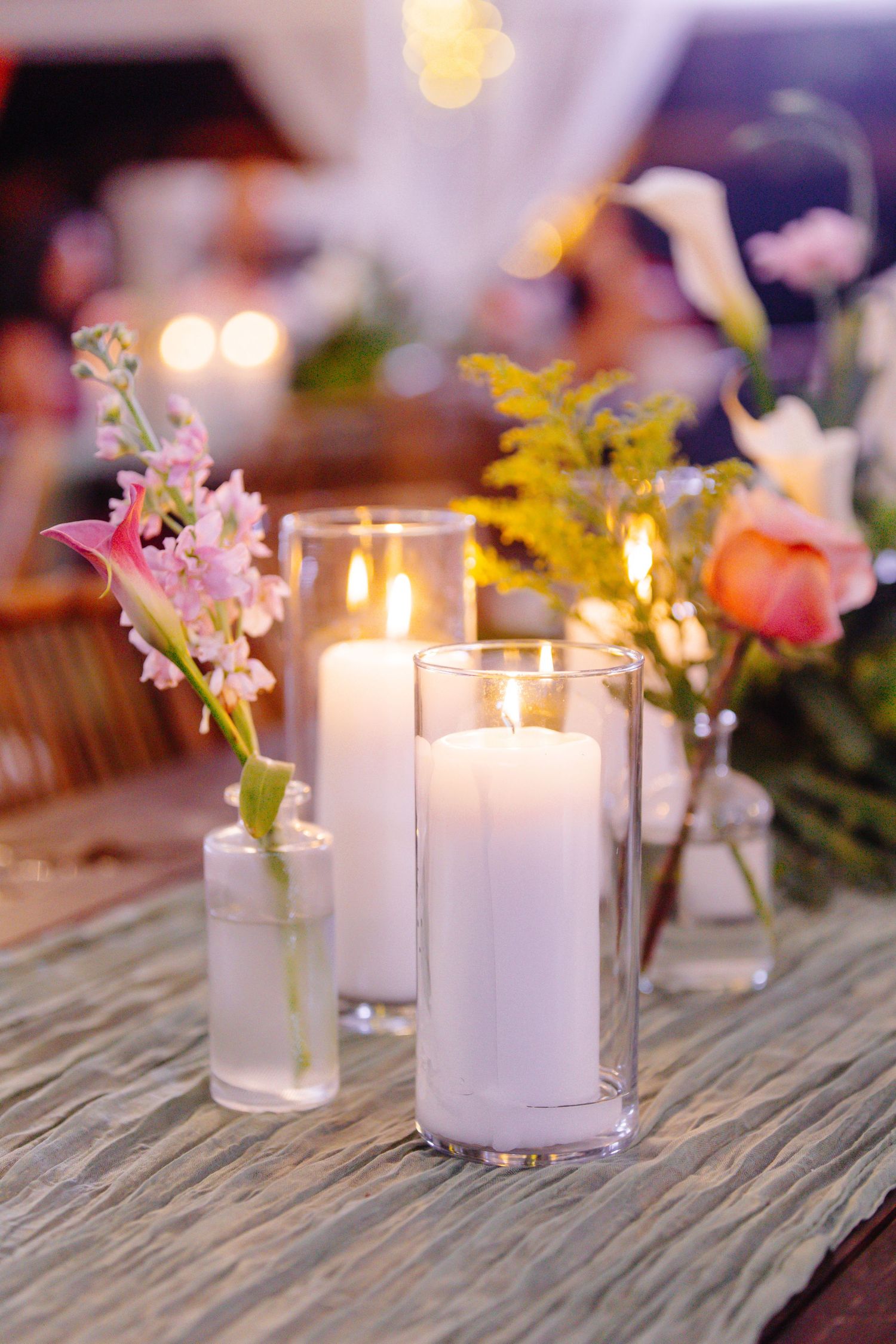 Elegant wedding table centerpiece with white pillar candles in glass holders surrounded by colorful fresh flowers on a rustic table.