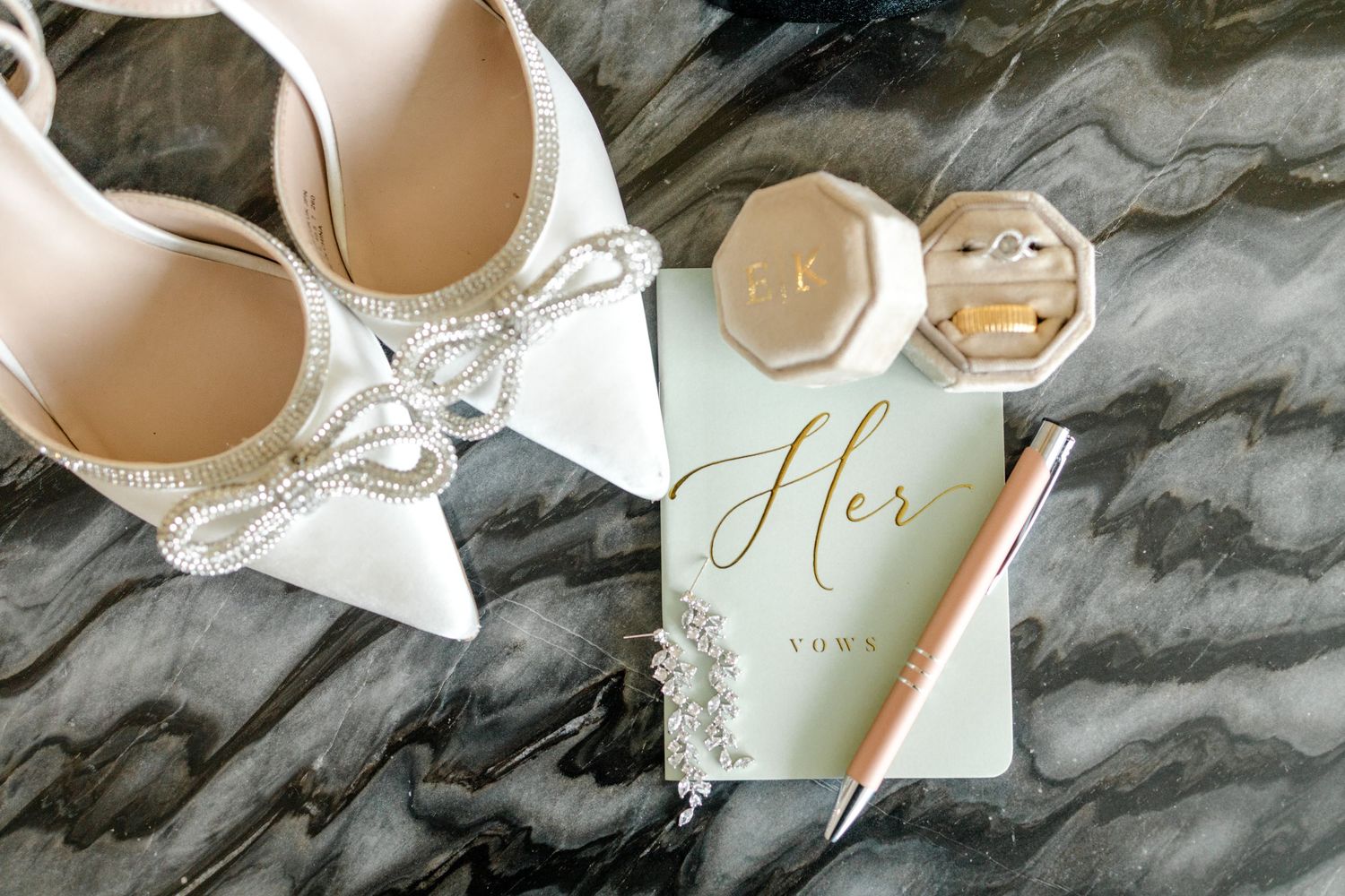 Bridal flat lay with white bow heels, vow book, ring box, and pearl earrings on marble surface.