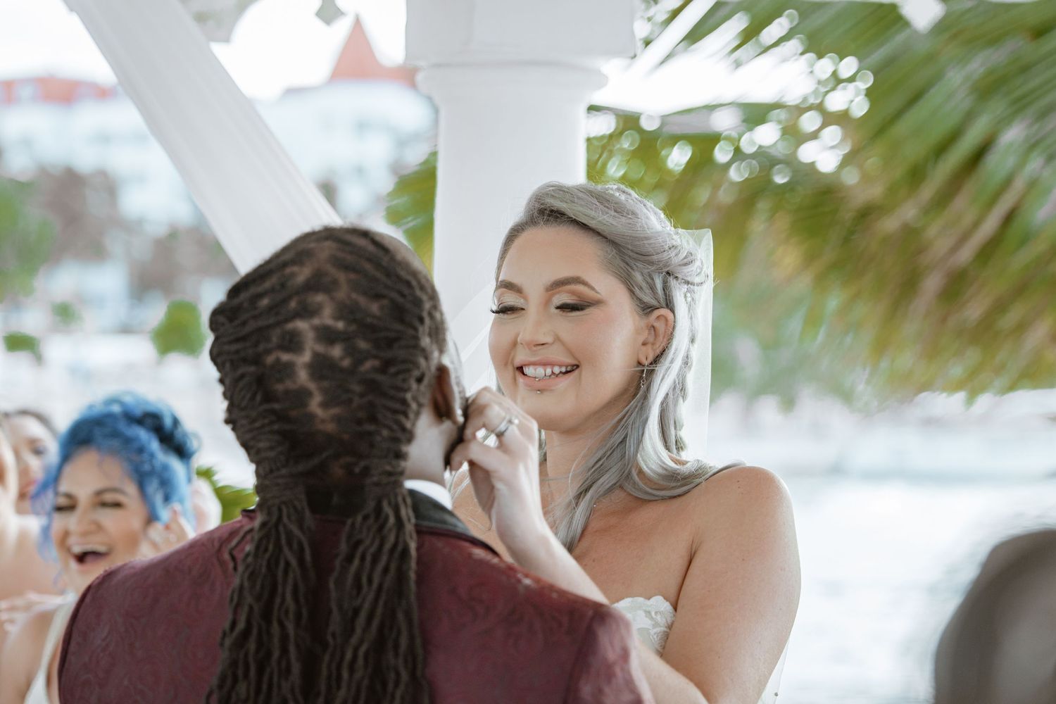 Smiling bride in white strapless gown exchanges rings with groom at sunlit wedding ceremony.