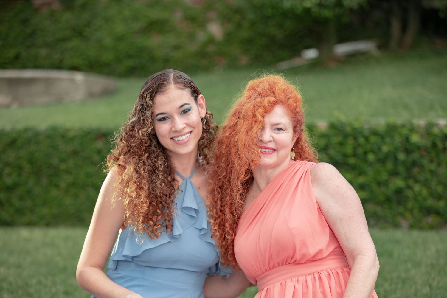 Two women with curly hair posing together outdoors in blue and coral dresses.