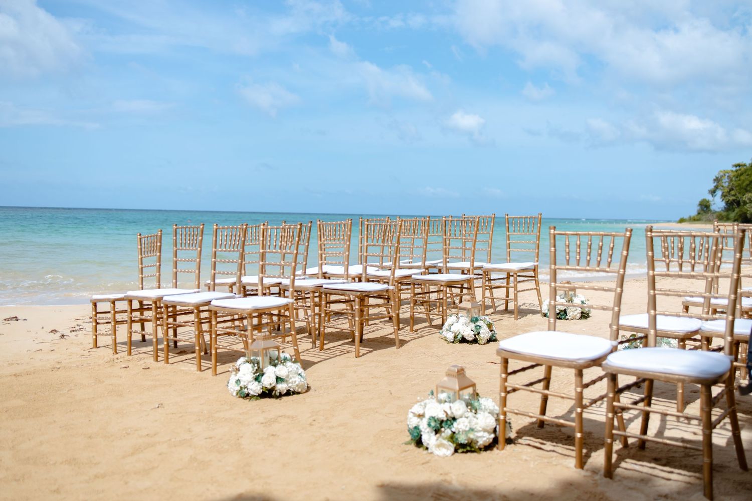 Gold chiavari chairs lined up on a sunny tropical beach for an outdoor wedding ceremony setup.