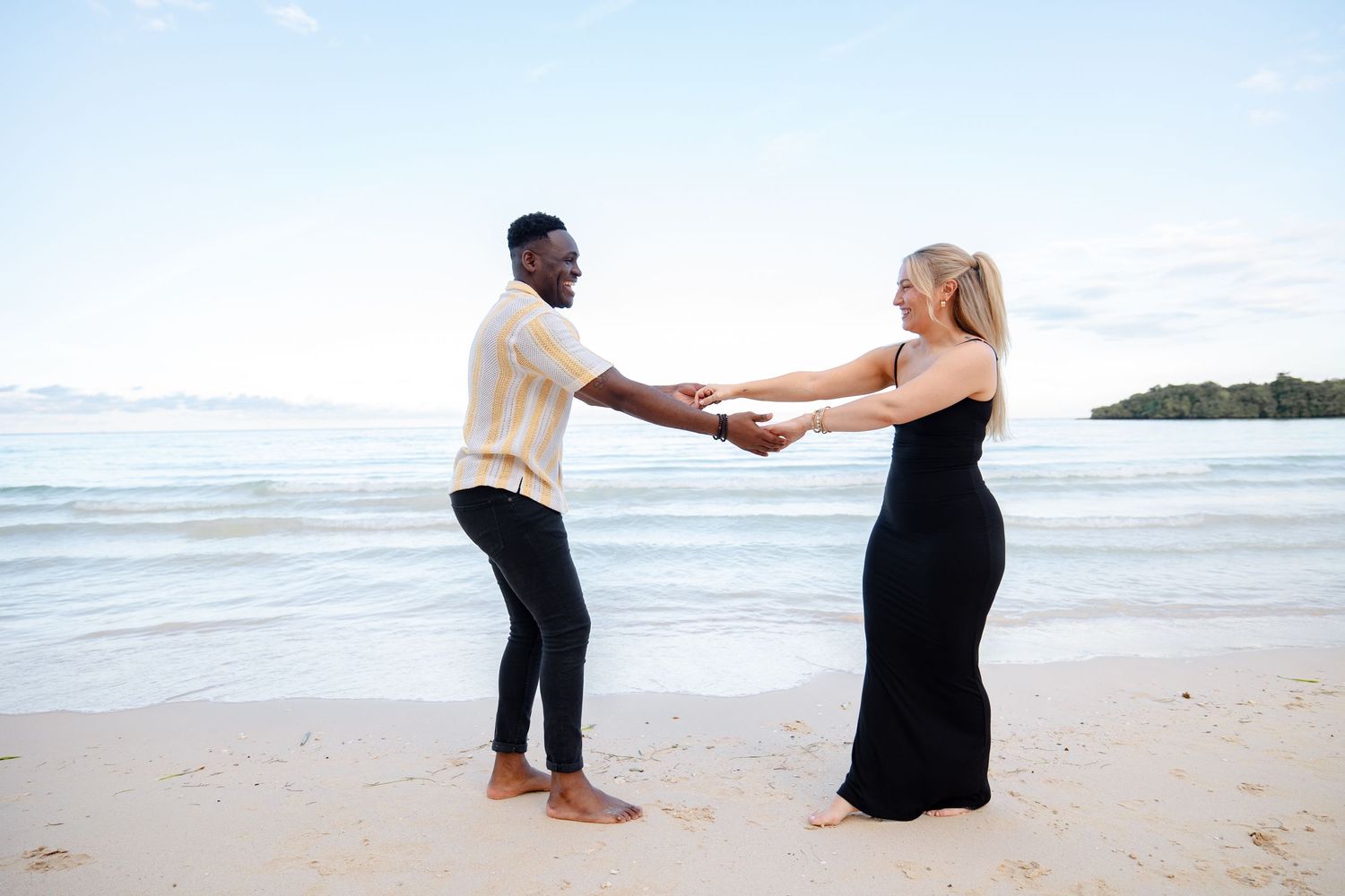 Interracial couple holding hands and laughing together on a sunny tropical beach shoreline.
