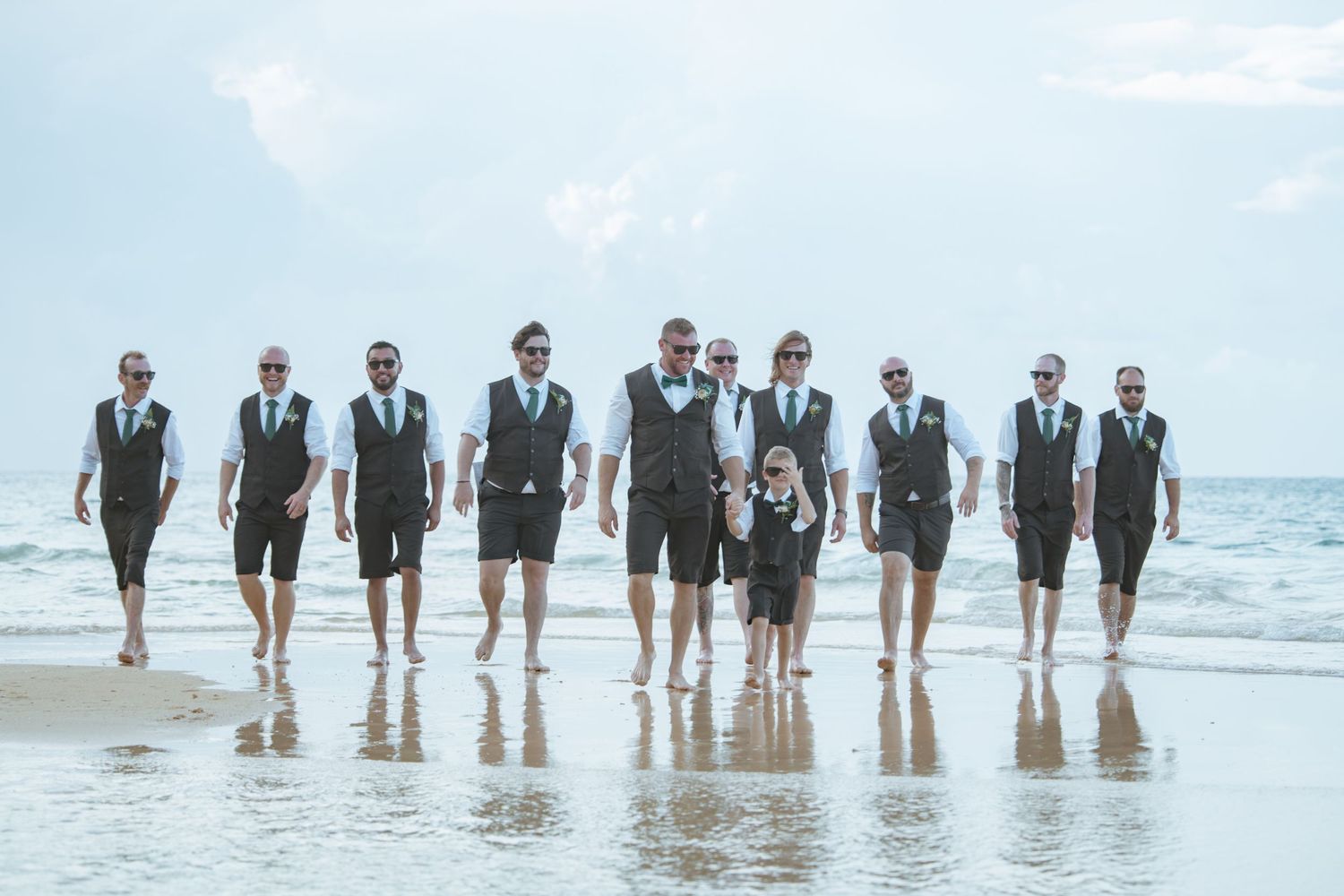 Groomsmen in matching dark vests and shorts walking confidently along a beach shoreline.