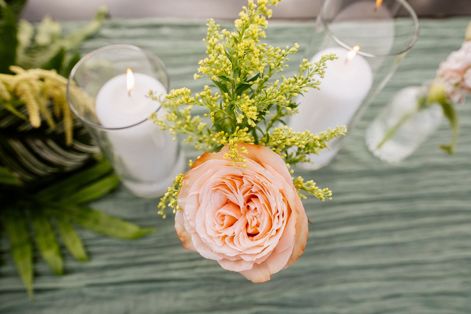 Peach garden rose and yellow mimosa flowers beside white pillar candles in glass holders on a green table runner.