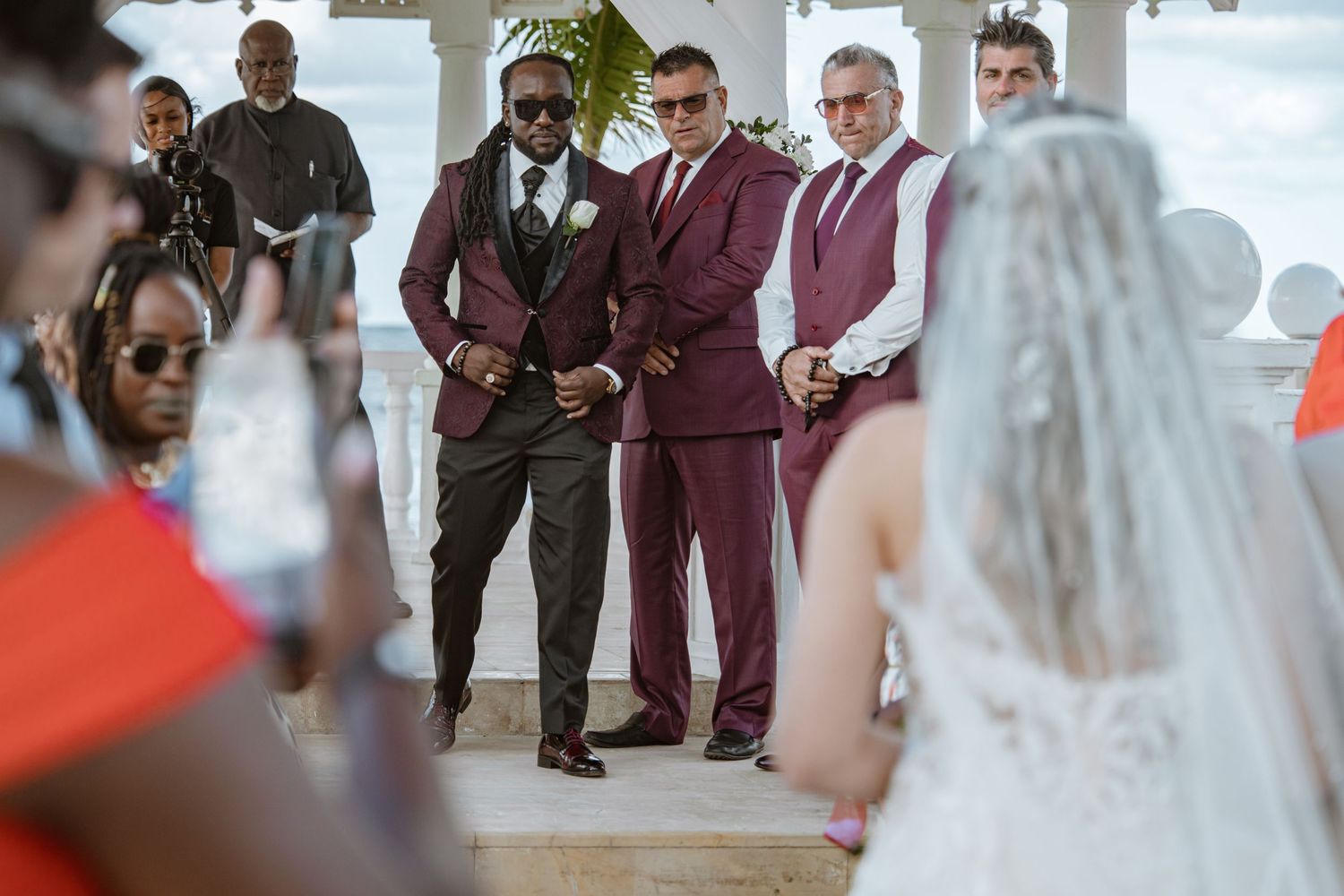 Groom in burgundy suit stands at altar with groomsmen as bride approaches during wedding ceremony.