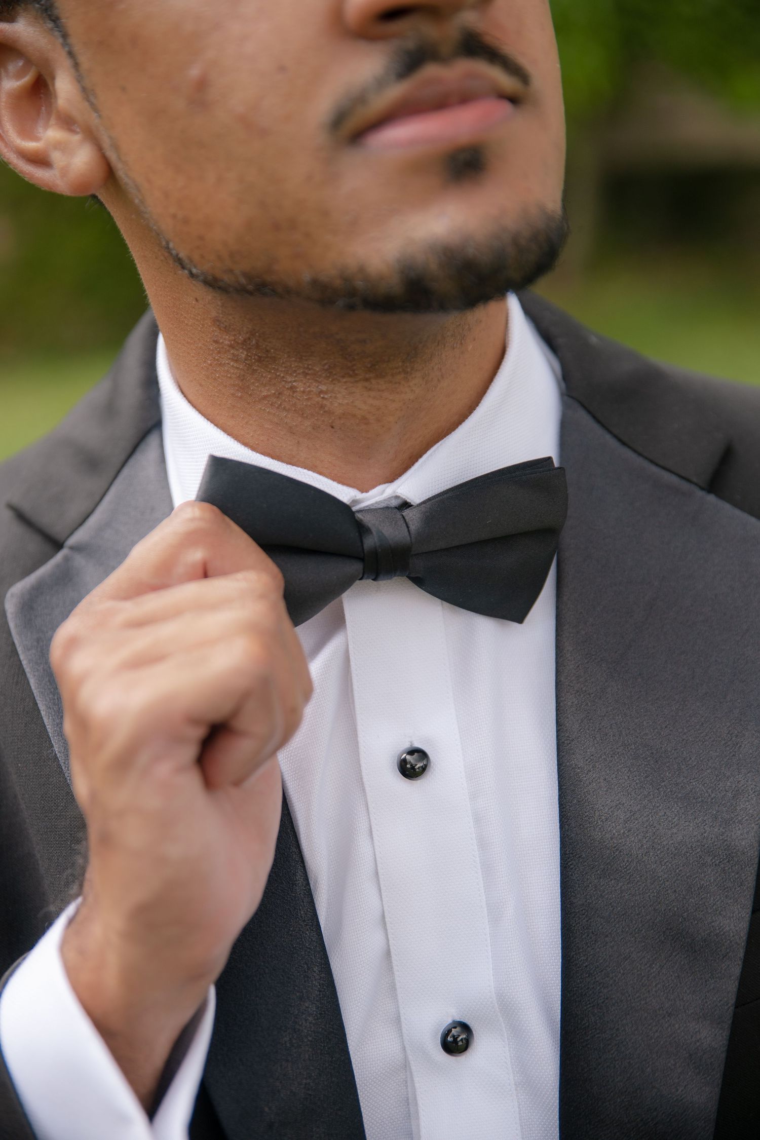 Groom in black tuxedo adjusting bow tie outdoors with green foliage background.