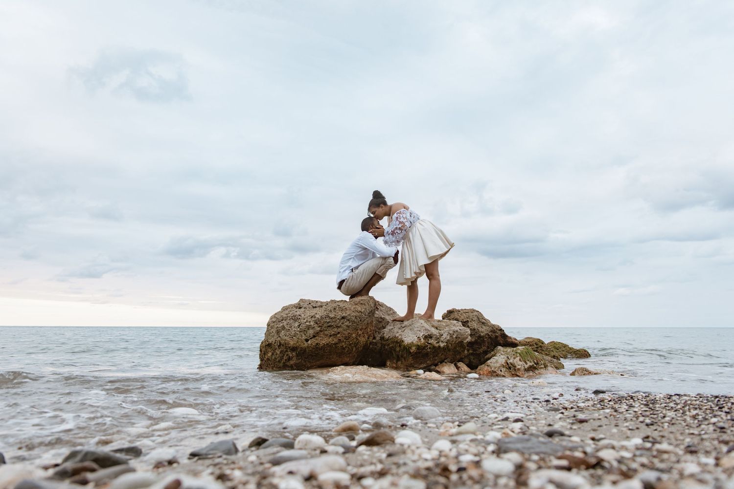 Romantic couple sharing a tender kiss while standing on a rock by the serene seaside under a cloudy sky.