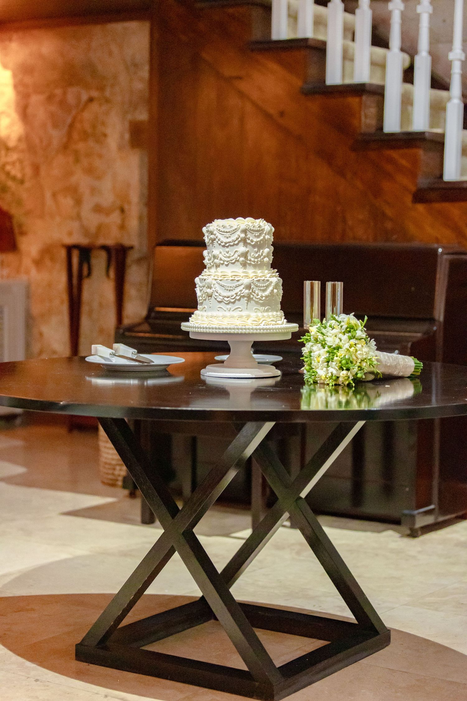 Elegant three-tier white ruffled wedding cake displayed on dark wooden table beside bridal bouquet.