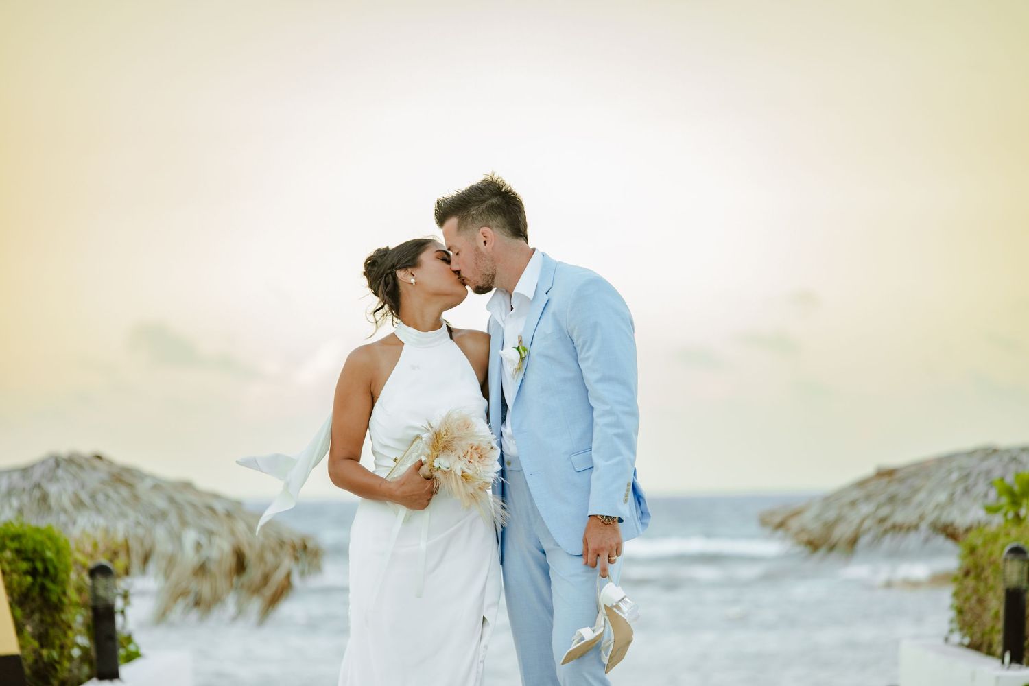 Couple sharing a romantic kiss at a beachside wedding, bride in white dress, groom in light blue suit.