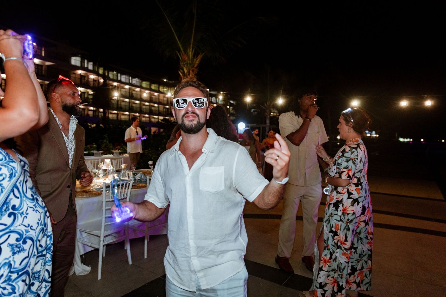 Guest in white outfit and novelty glasses dancing at outdoor beach wedding reception at night.