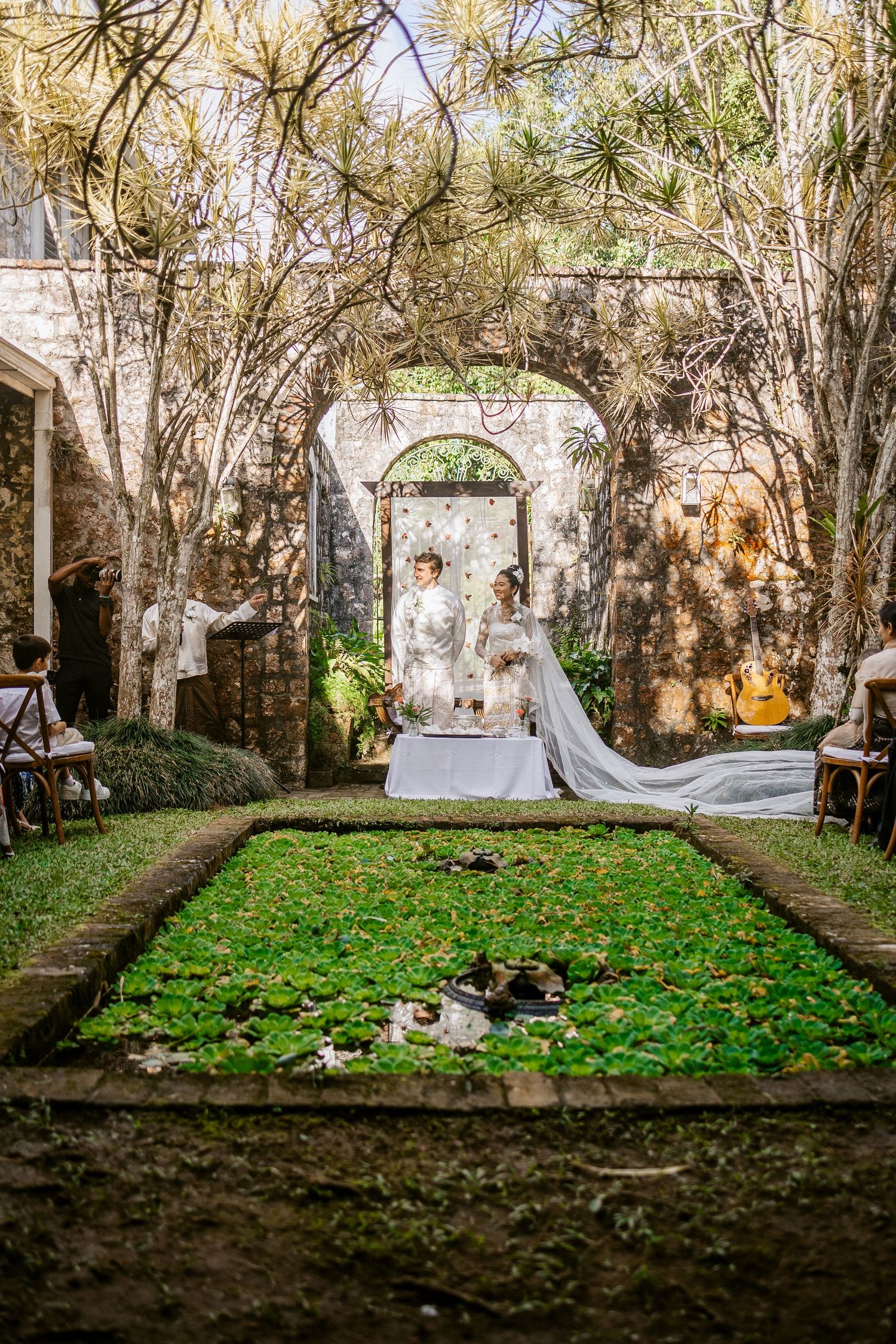 Elegant outdoor wedding ceremony in a lush garden courtyard with stone arches, bride and groom at altar.