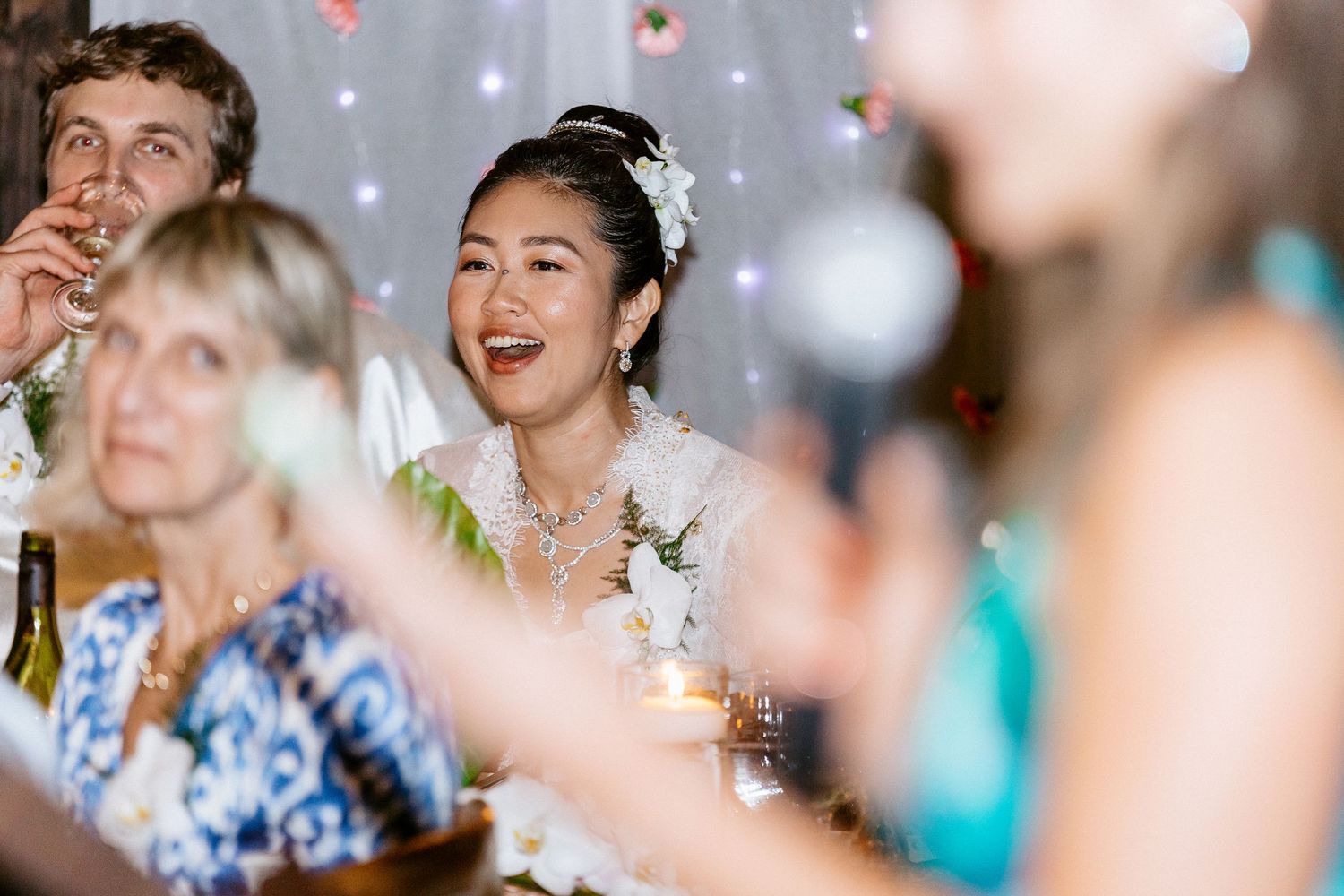 Joyful bride in white floral headpiece laughing during wedding reception with guests.