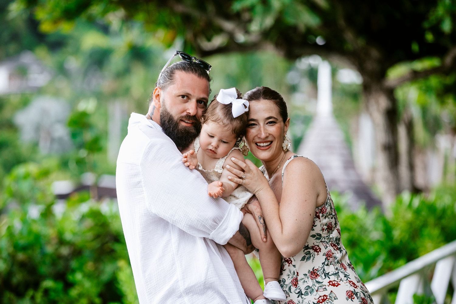 Happy family with bearded father, smiling mother in floral dress, and baby girl with white bow posing outdoors.