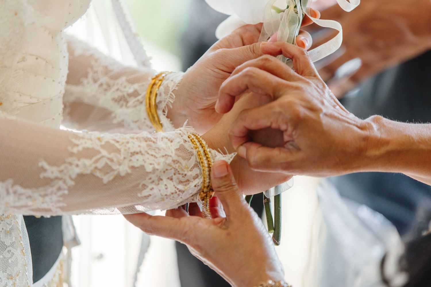 Close-up of hands exchanging gold bracelets during a traditional wedding ceremony, bride wearing white lace dress.