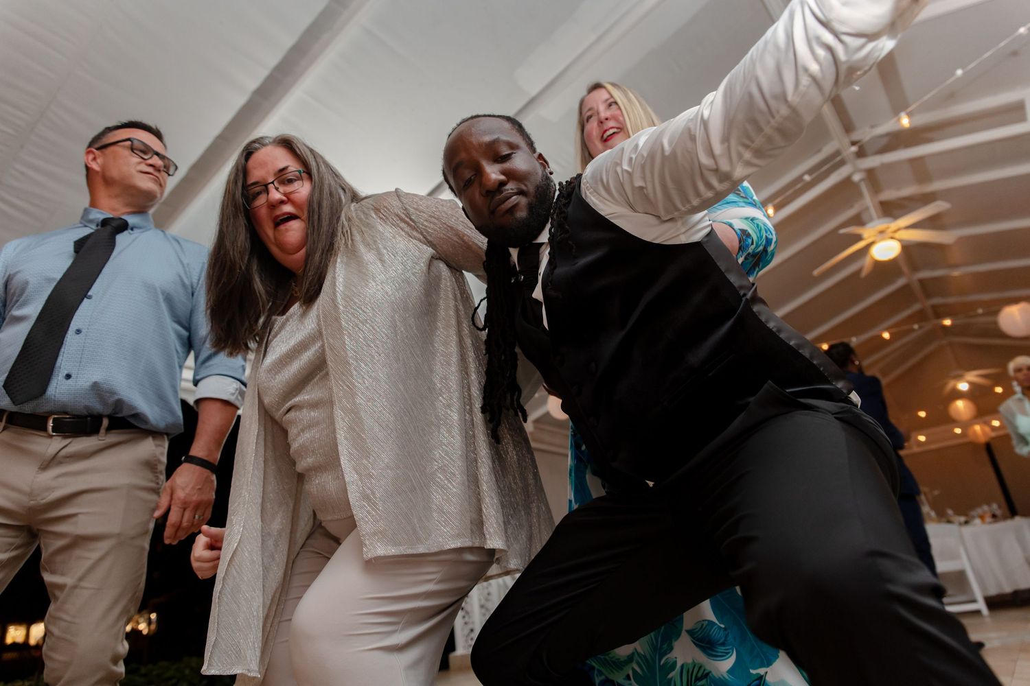 Energetic wedding guests dancing together under white tent with string lights at evening reception.