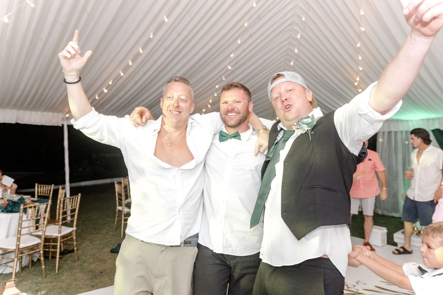 Groom celebrating joyfully with two friends at an outdoor tented wedding reception.