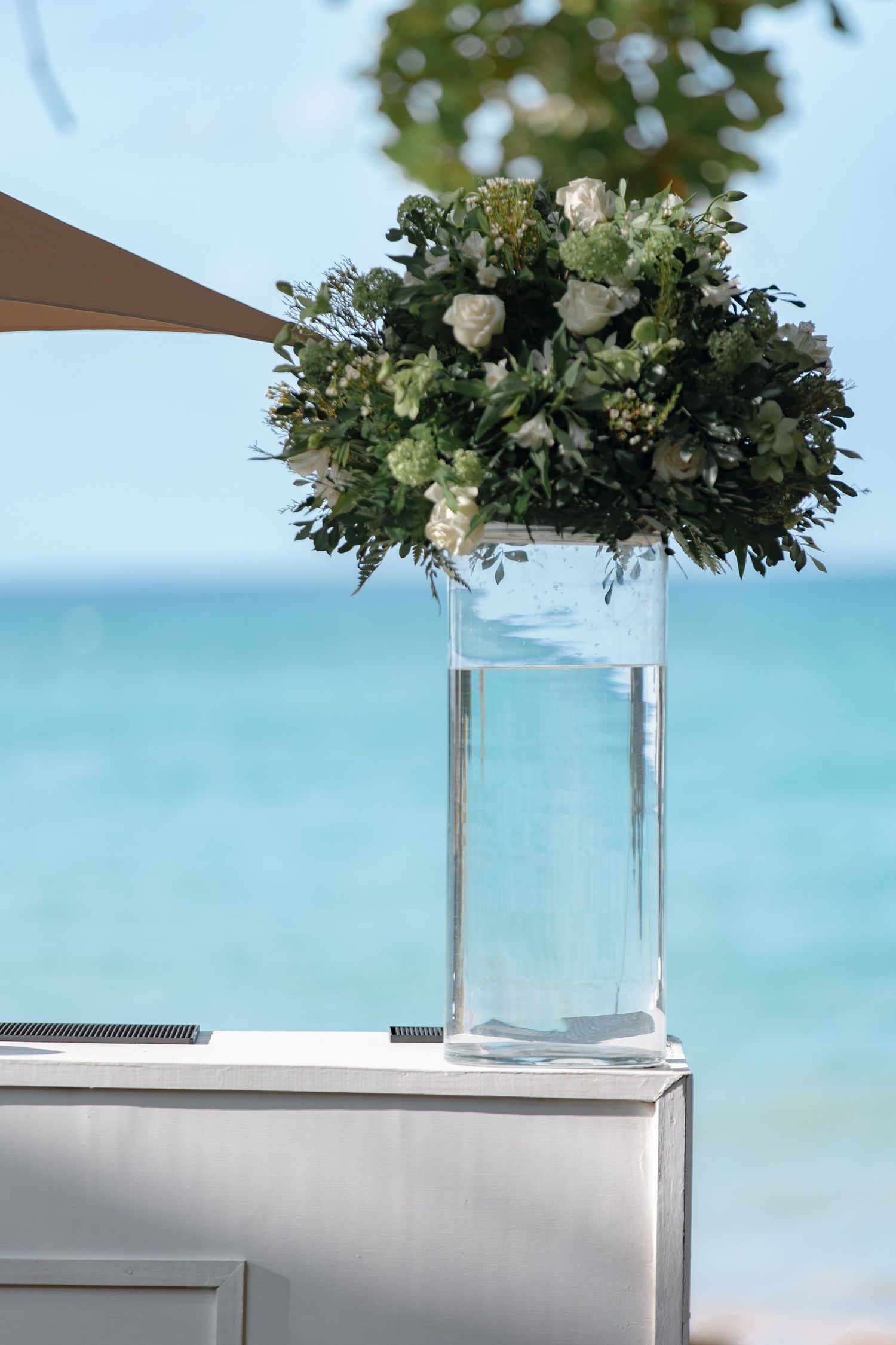 White floral arrangement in tall glass vase on white pedestal with turquoise ocean in background.