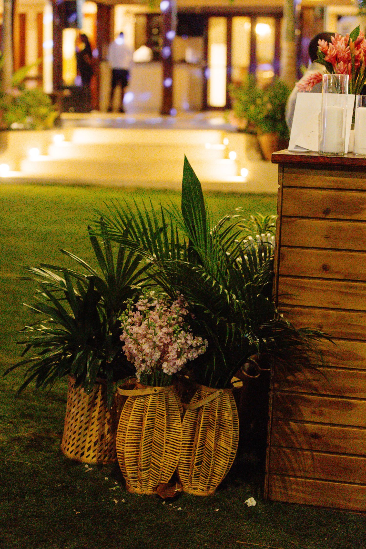 Wicker baskets with tropical palm plants and pink flowers beside a wooden bar at an outdoor evening event.