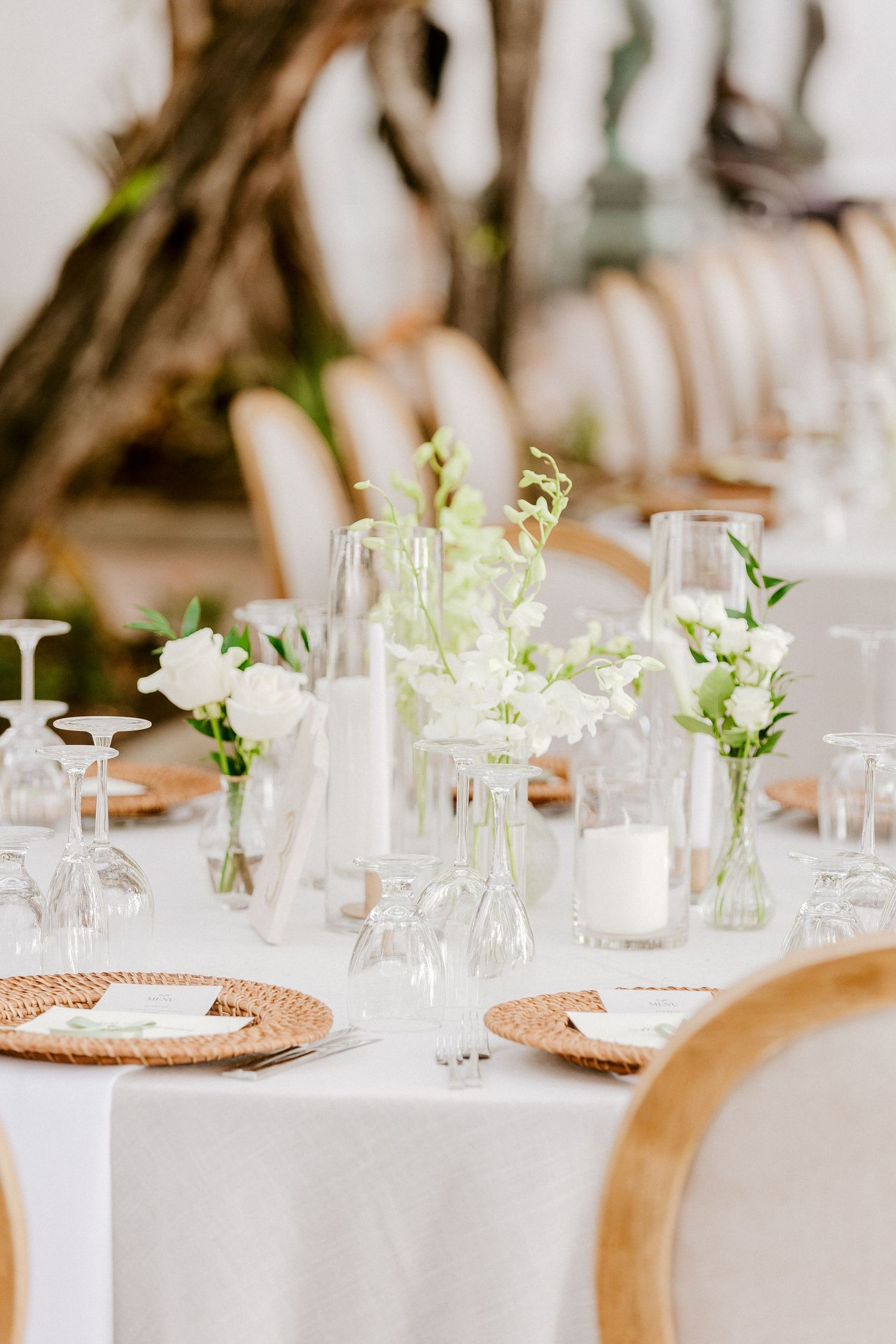 Elegant white wedding table setting with floral arrangements, glass vases, candles, and woven charger plates.