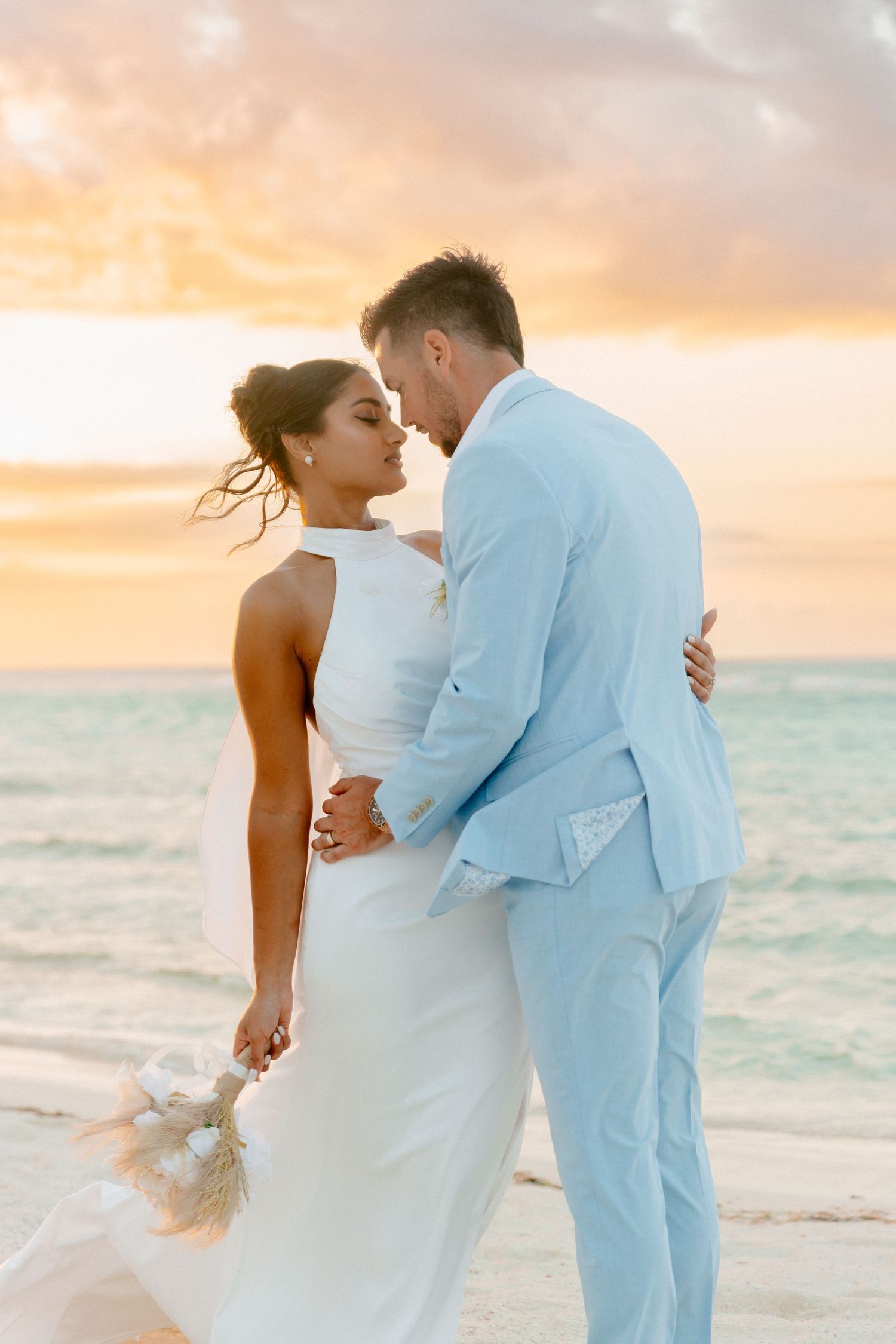 Bride and groom share a romantic moment on the beach at sunset, with turquoise ocean waves in the background.