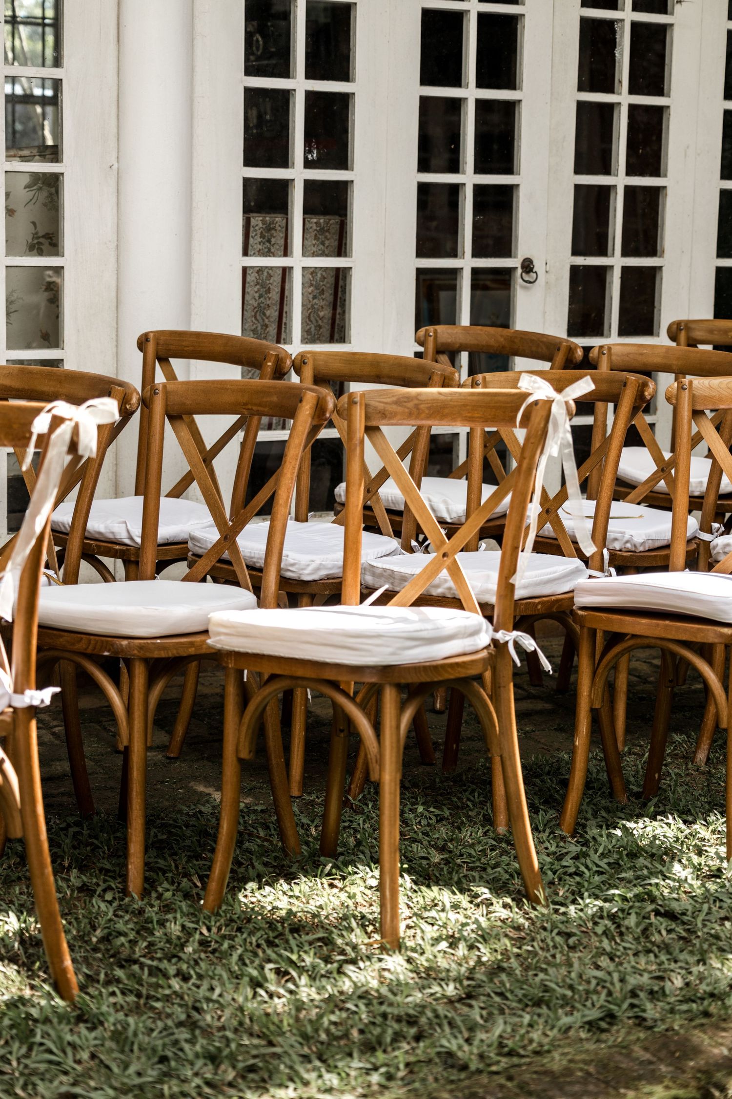 Rows of wooden cross-back chairs with white cushions and ribbon ties set up for an outdoor wedding ceremony.