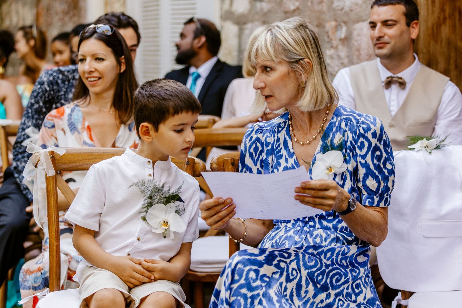Woman in blue floral dress reads a paper to a young boy in white at a sunny outdoor wedding ceremony.