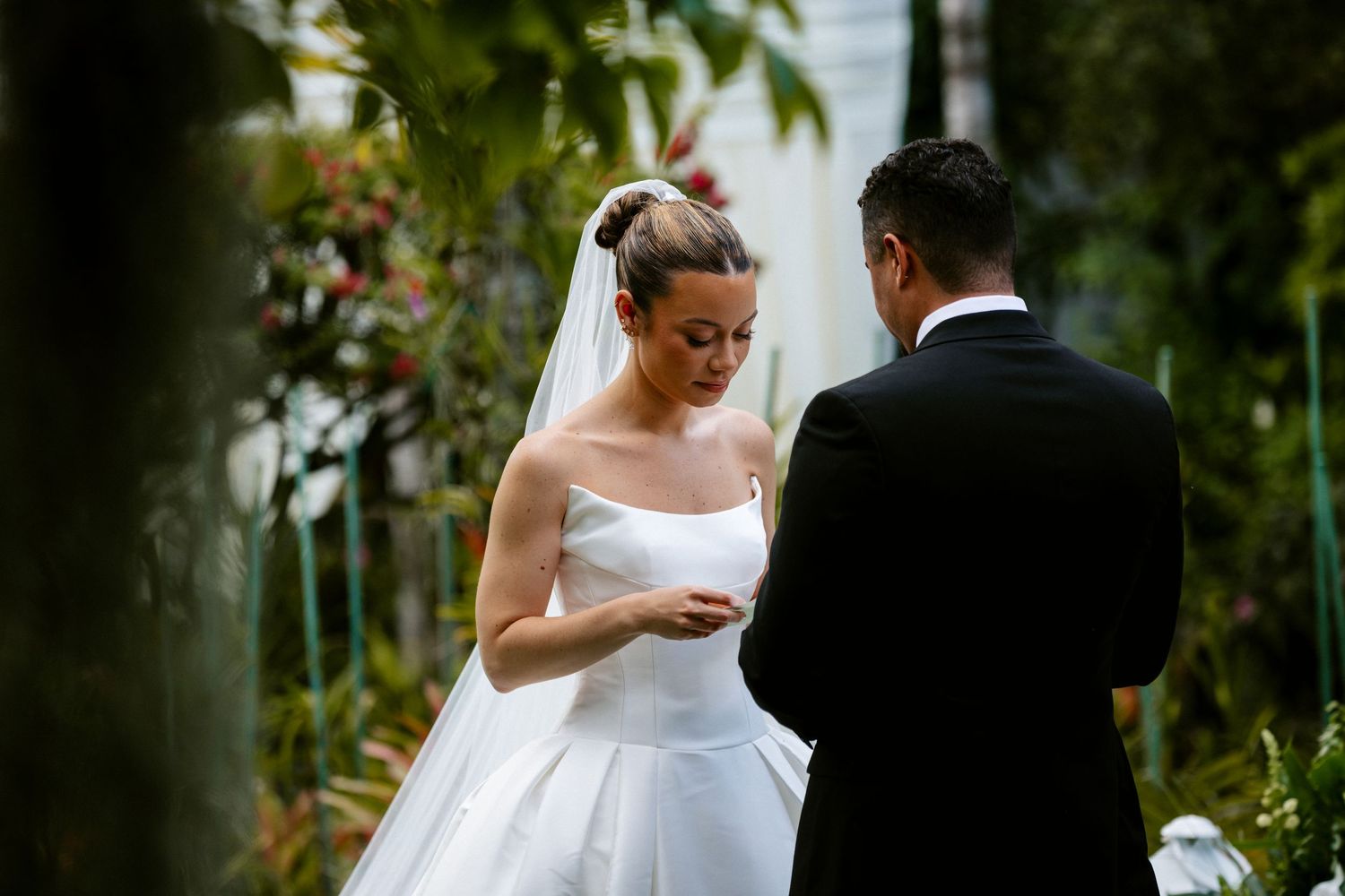 A bride in a strapless white wedding gown reads her vows during an outdoor garden ceremony with her groom.