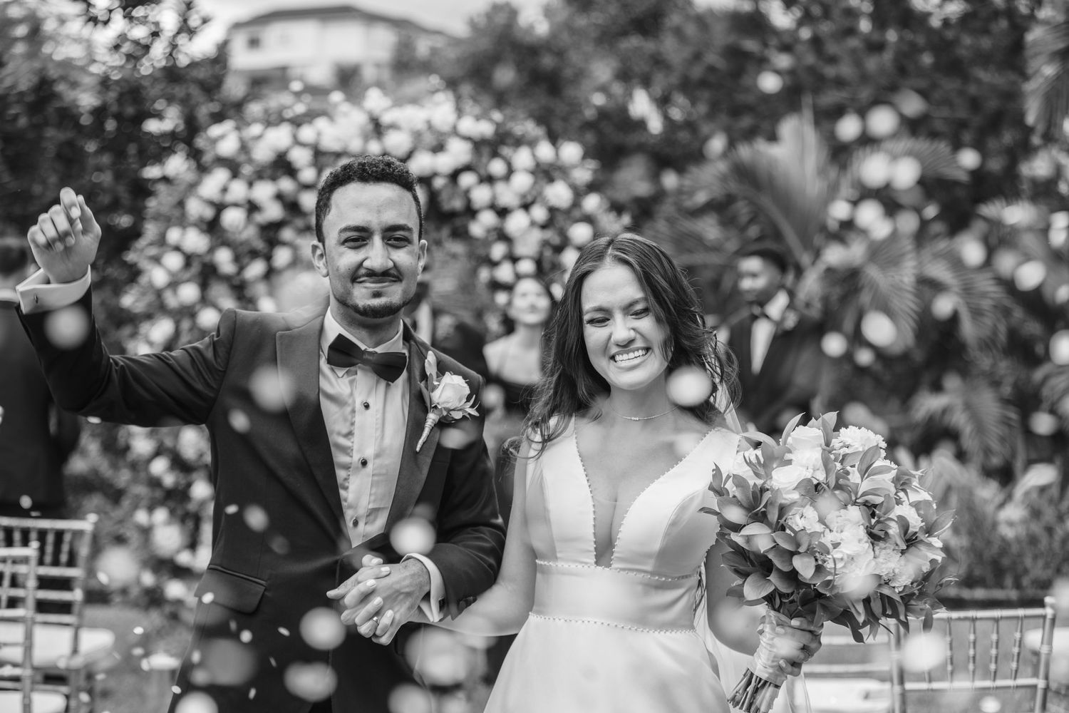 Black and white photo of happy newlyweds walking through confetti shower at outdoor wedding.