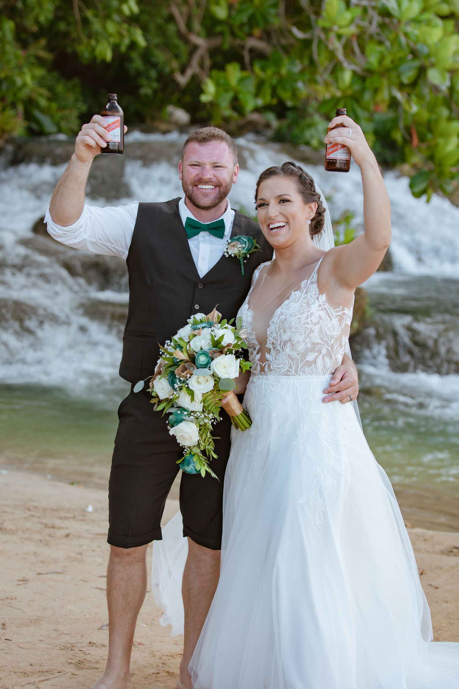 Smiling bride and groom toasting with beer bottles near a tropical waterfall on beach.