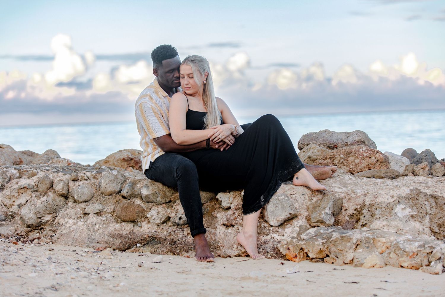 Interracial couple sitting closely together on rocky beach wall with ocean and clouds behind them.