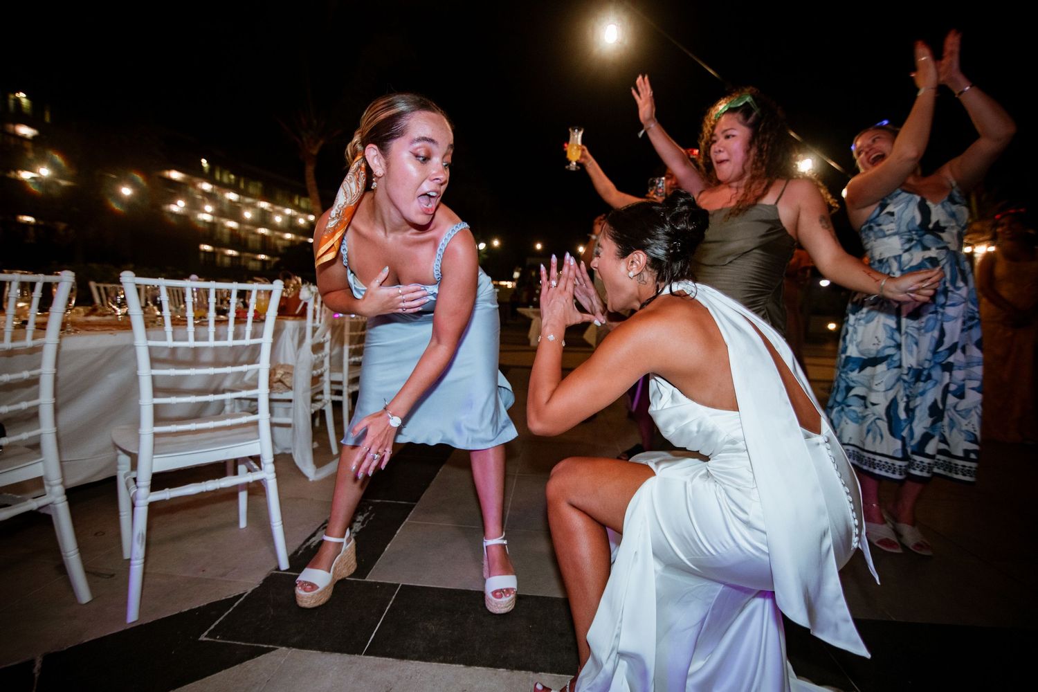 Excited women celebrating at a wedding reception outdoors at night, with white chairs and festive atmosphere.