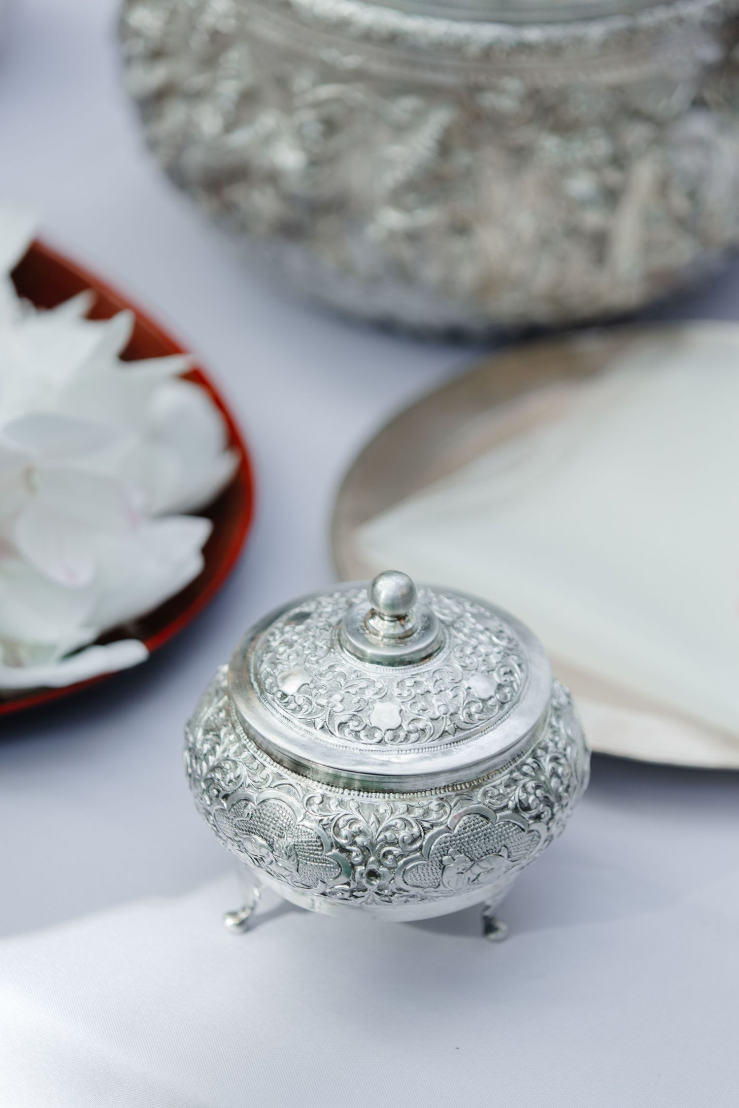 Ornate silver Thai ceremonial bowl with intricate floral engravings and lid, displayed on white cloth with white flowers.