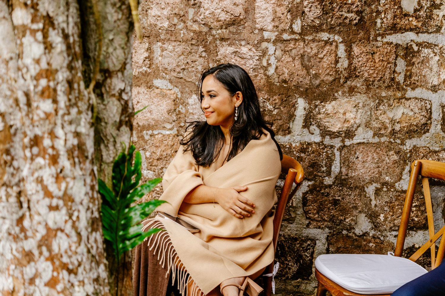 Woman wrapped in beige fringed shawl sitting thoughtfully against a rustic stone wall outdoors.
