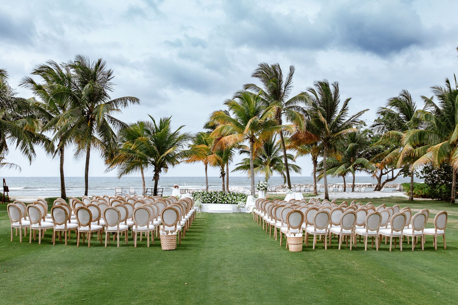 Outdoor wedding ceremony setup with white chairs on lush lawn surrounded by palm trees overlooking the ocean.