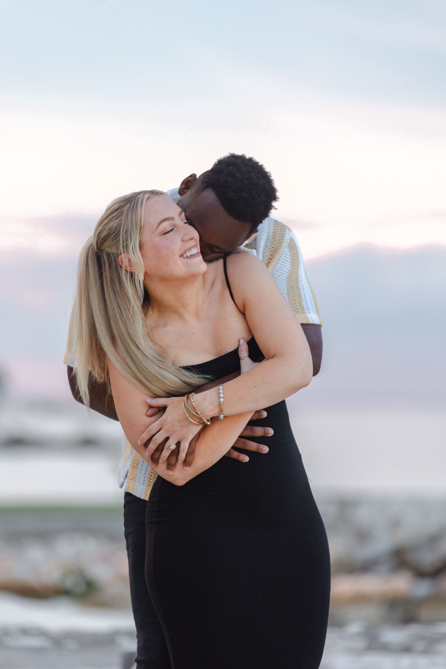 Interracial couple embracing on beach at sunset, man kissing womans neck as she laughs joyfully.