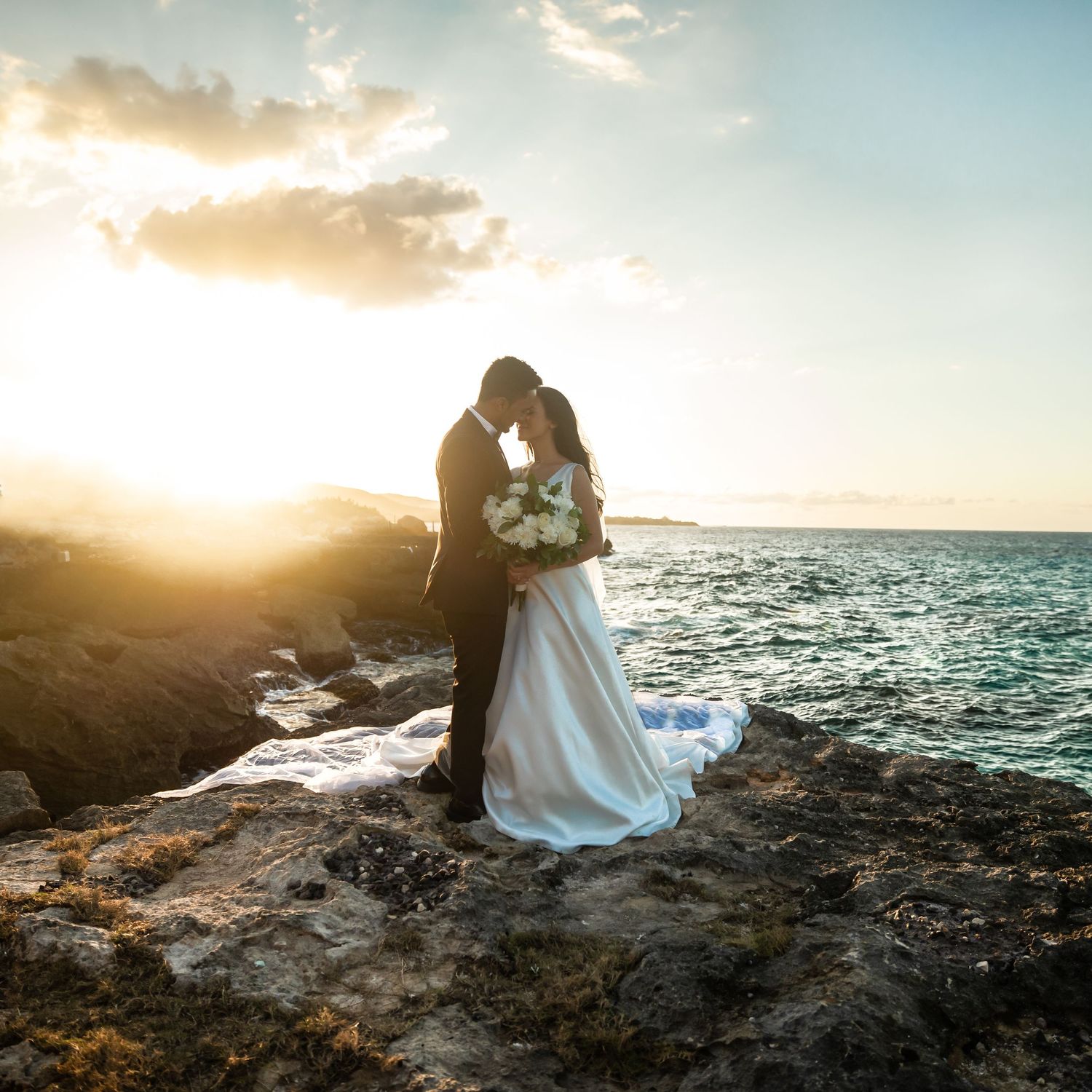 Bride and groom share romantic sunset kiss on rocky ocean cliff, wedding veil flowing in wind.