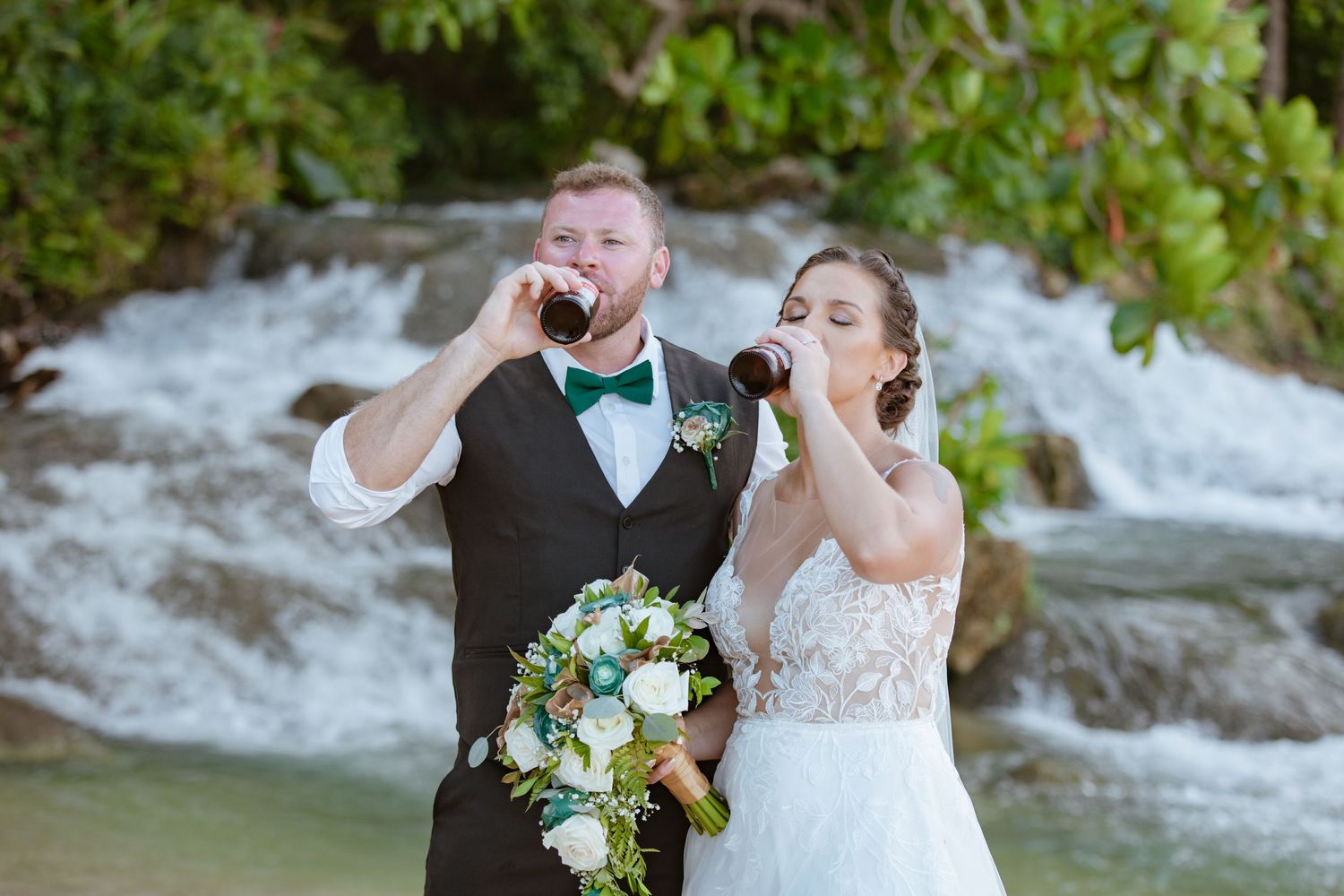 Bride and groom drinking beer bottles together near a tropical waterfall on their wedding day.