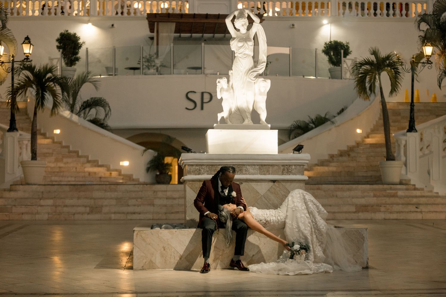 Newlyweds pose dramatically at night by a lit fountain statue at a resort wedding venue.