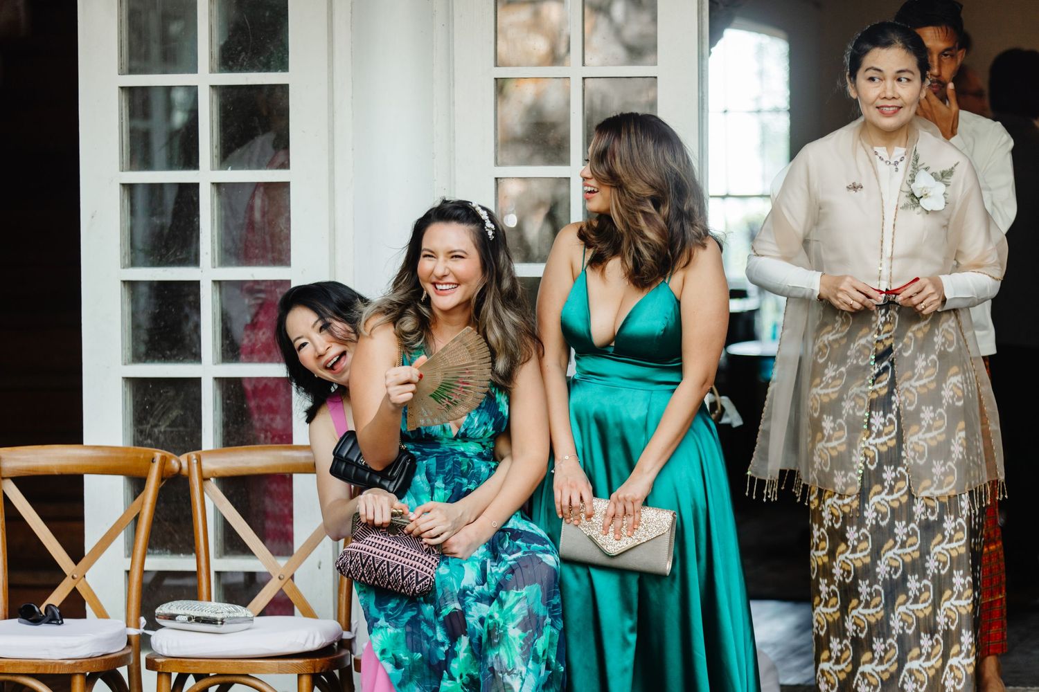 Four women in elegant formal attire laughing and socializing at an outdoor wedding reception.