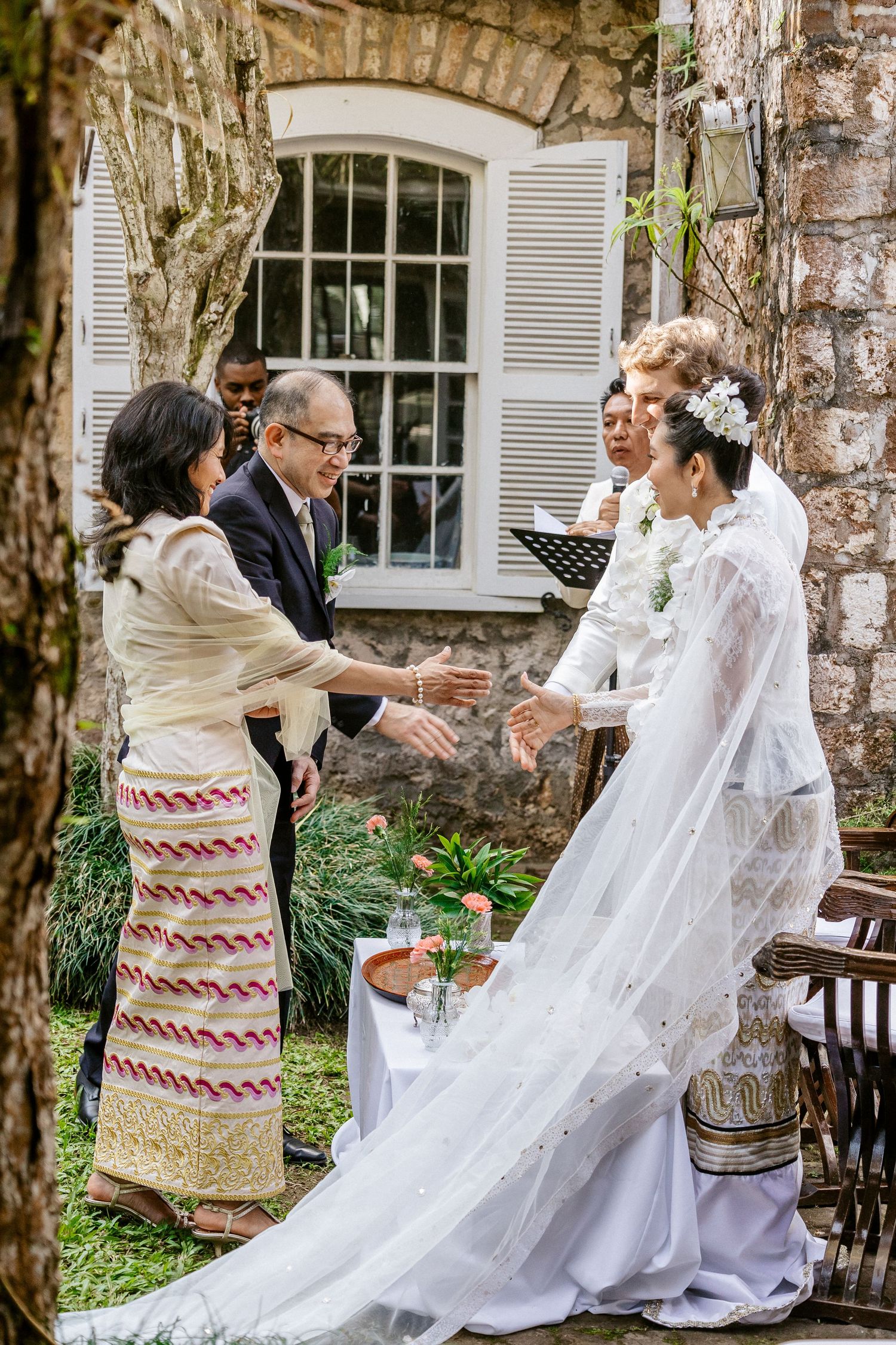 Outdoor wedding ceremony with bride in white gown and veil greeting guests near stone wall and lush greenery.
