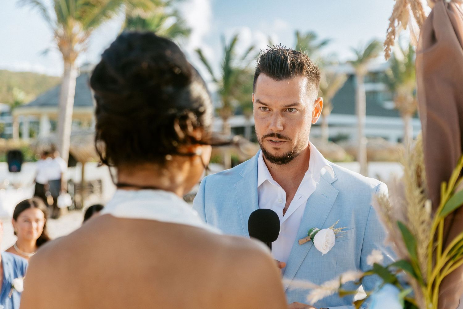 Groom in light blue suit listening attentively during outdoor beach wedding ceremony with tropical backdrop.