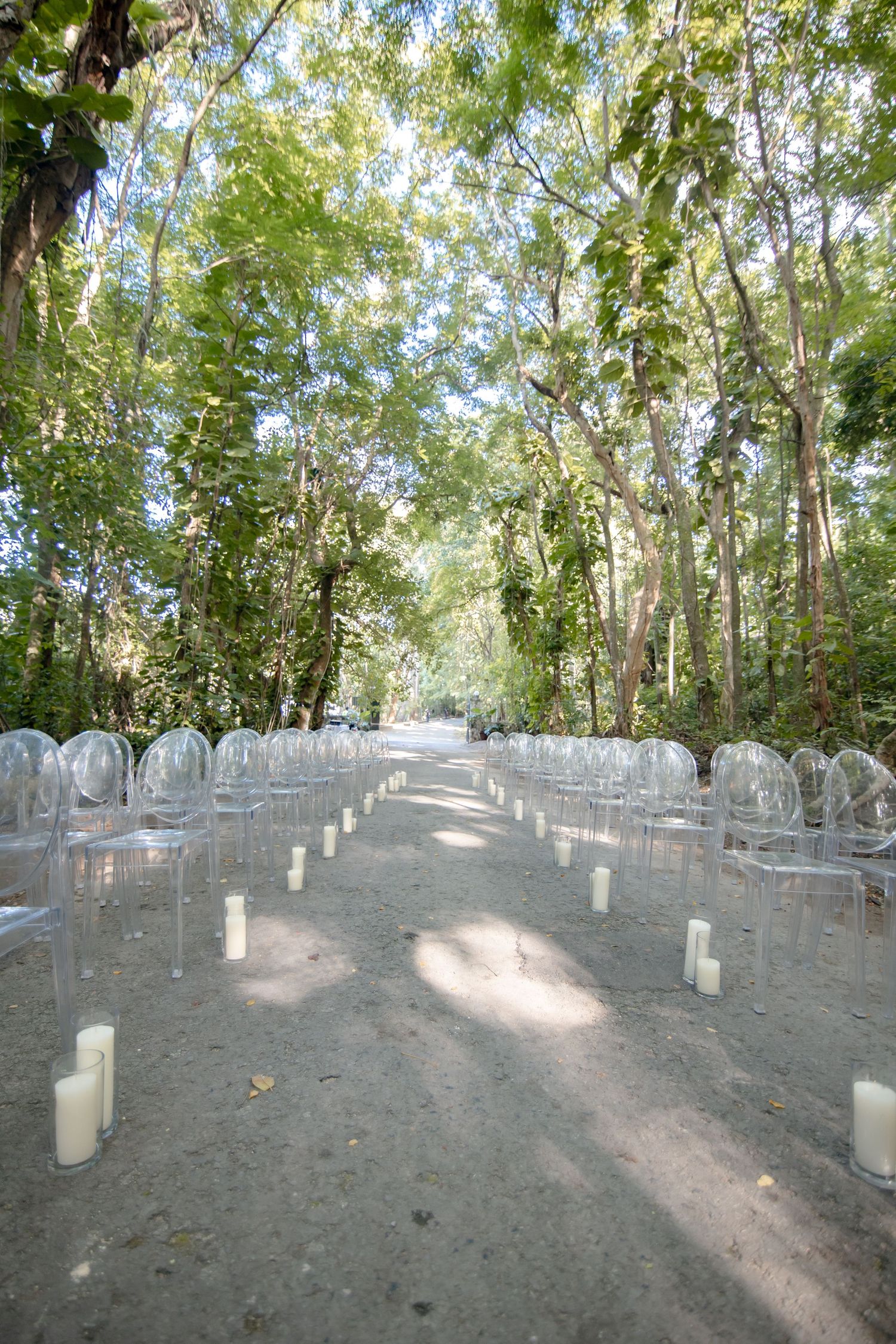 Elegant outdoor wedding ceremony aisle with transparent ghost chairs and white pillar candles lining a forest path.