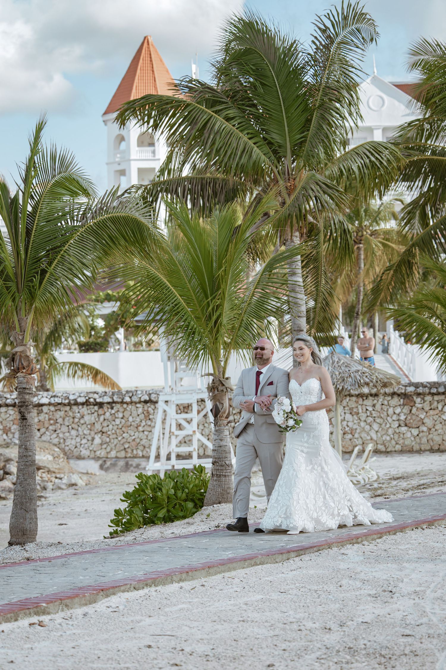 Bride and groom walking along a tropical beach path lined with palm trees at a destination wedding resort.