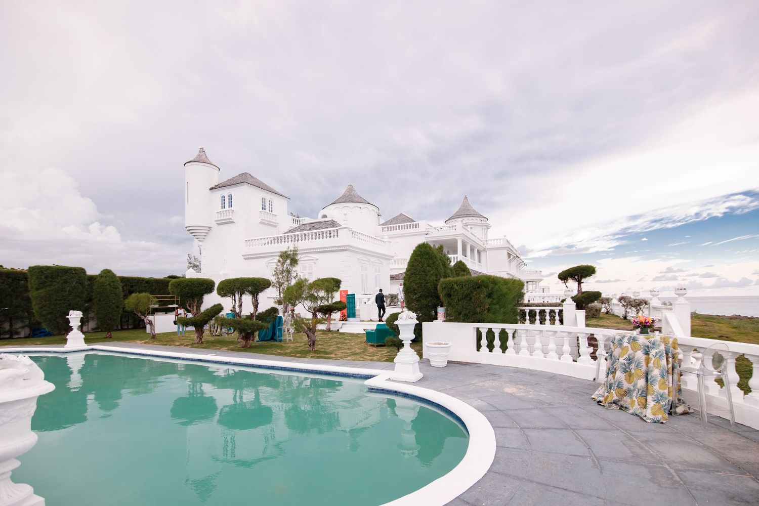 Elegant white mansion with turrets overlooks a turquoise pool, guests mingling on terrace under cloudy sky.