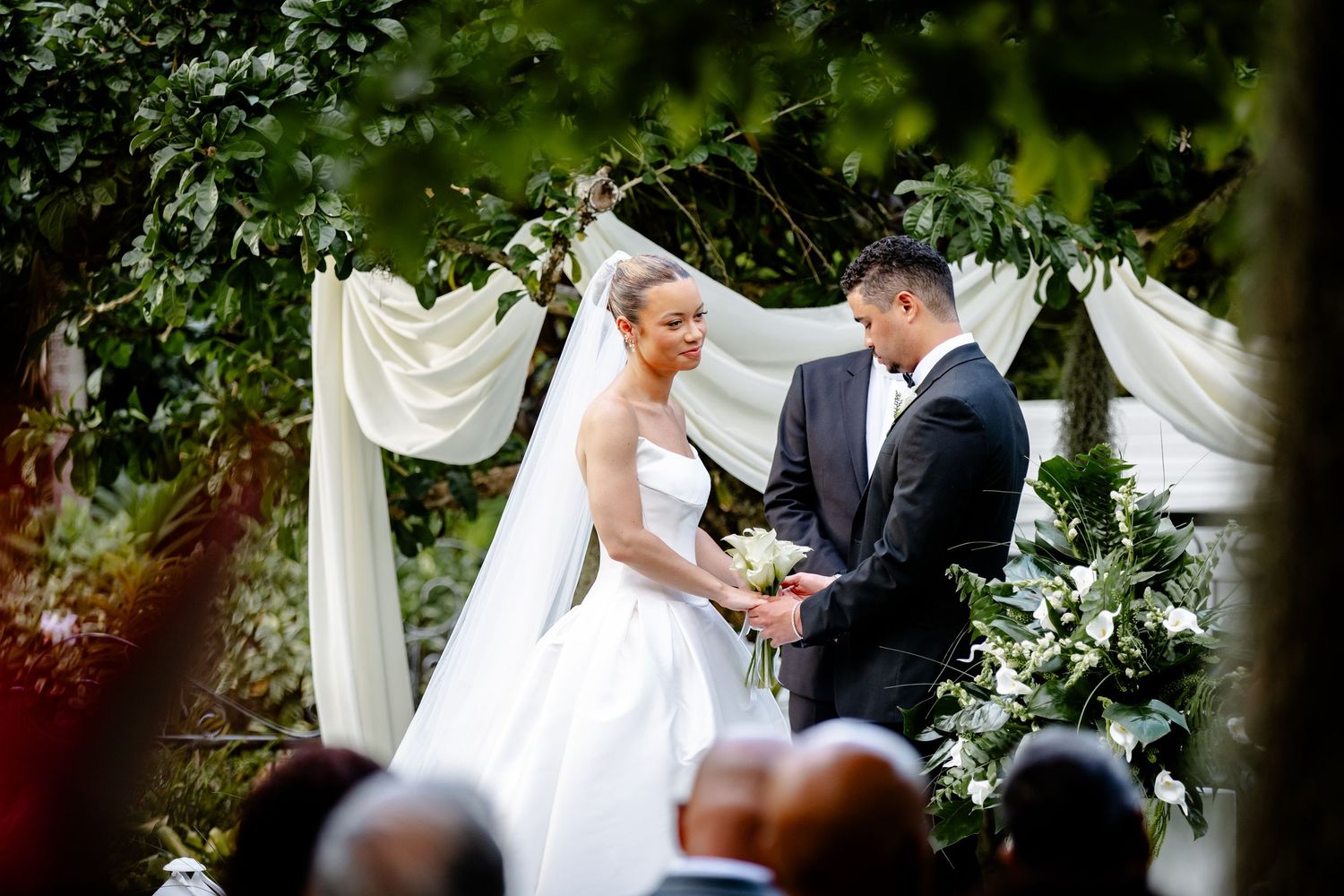 Bride and groom exchanging vows at an elegant outdoor ceremony beneath a white draped floral arch.