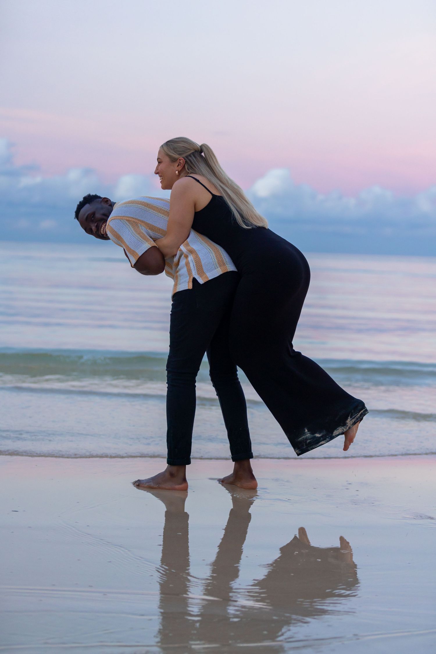 Couple sharing a joyful moment on a beach at sunset, man lifting woman near ocean waves.