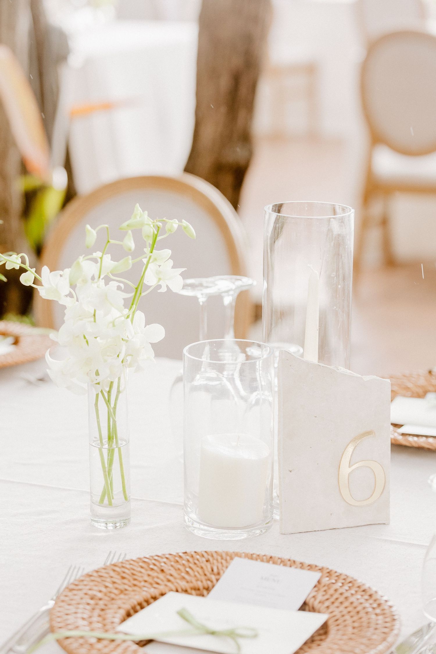 Wedding table centerpiece featuring white orchids, pillar candle in glass cylinder, and gold number six place card.
