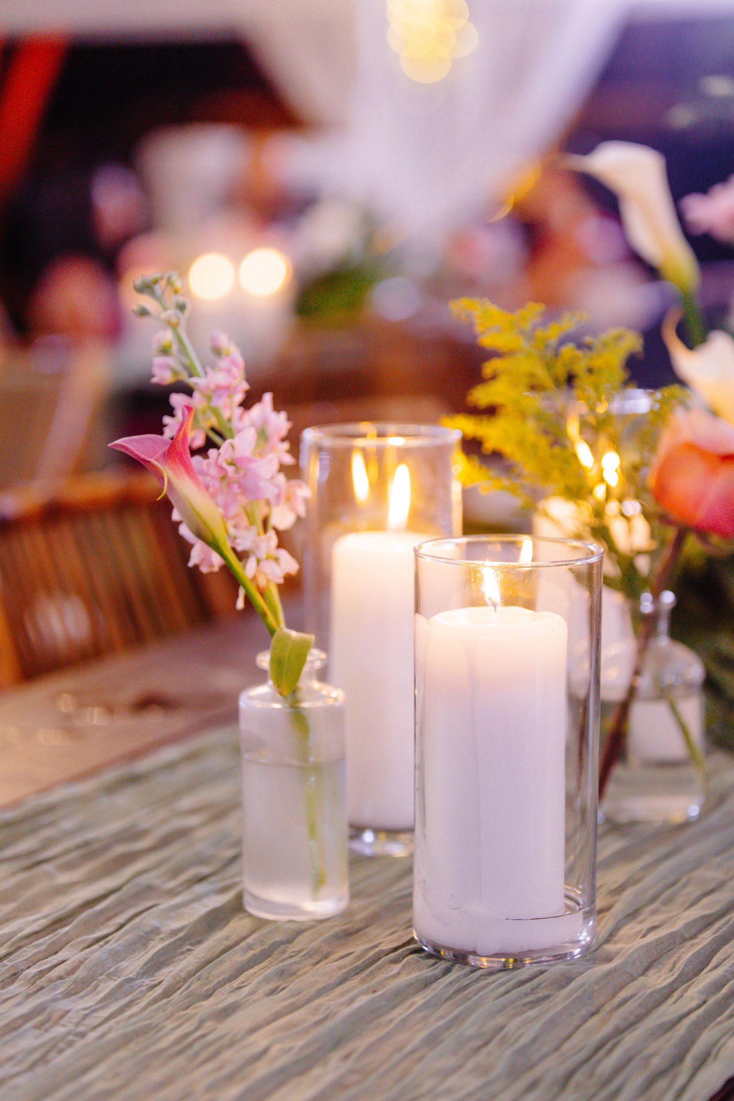 Elegant wedding table centerpiece with lit white pillar candles in glass holders and pink flowers in small bud vases.