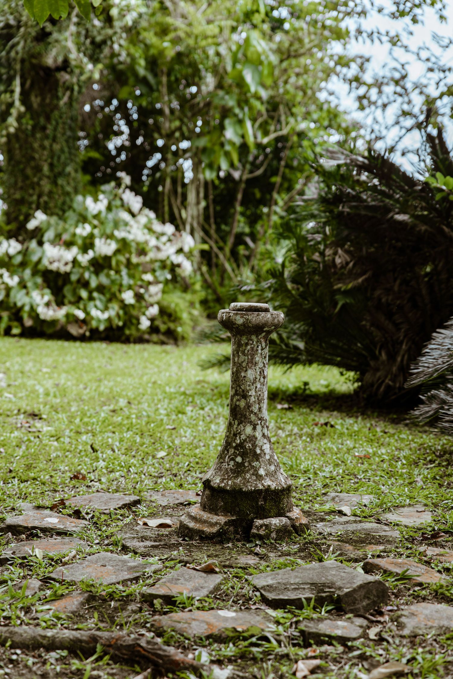 Ancient moss-covered stone sundial pedestal on cobblestone path in lush green garden with flowering trees.