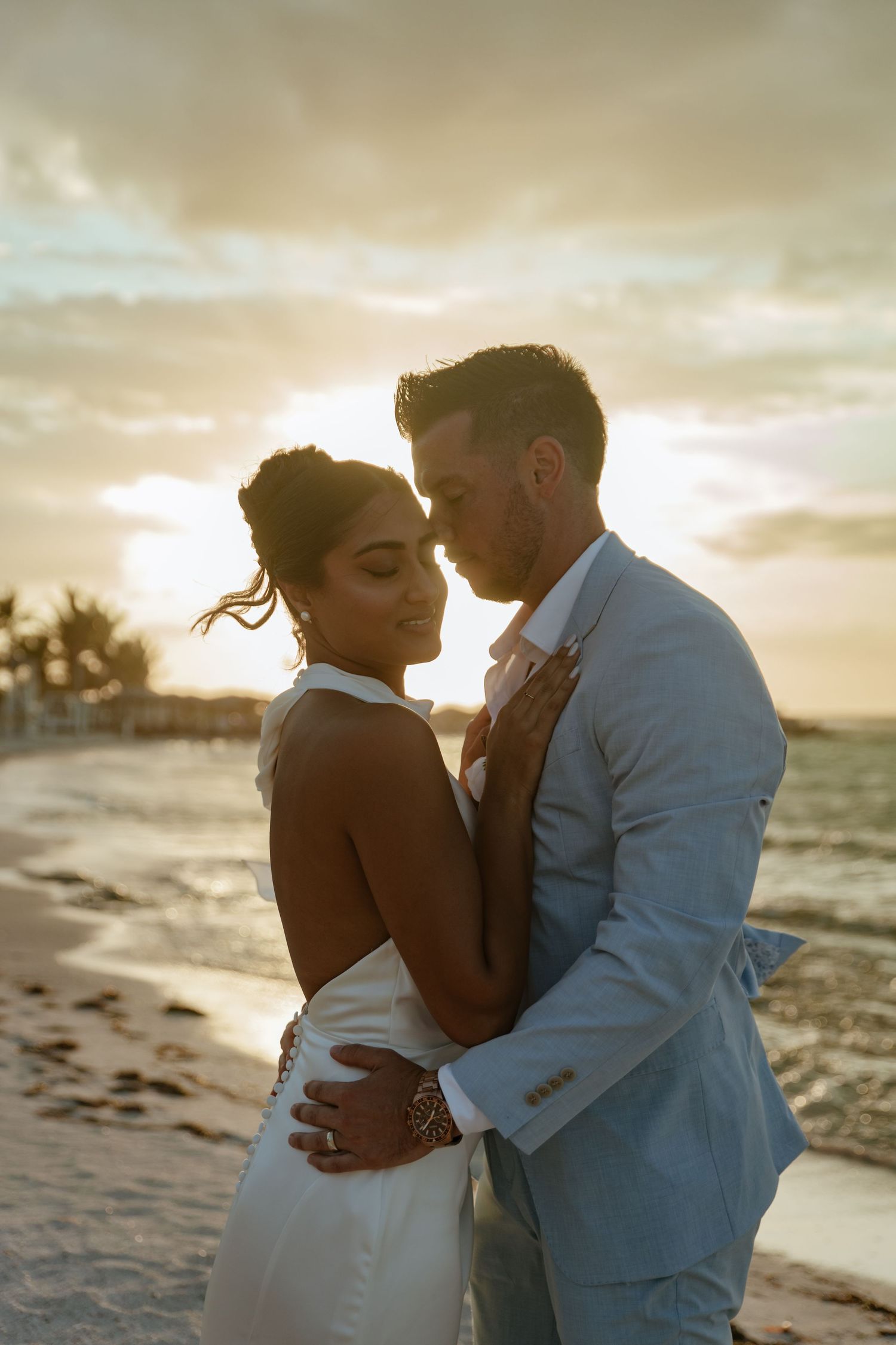 Bride and groom embracing romantically on beach at golden sunset in tropical destination wedding.